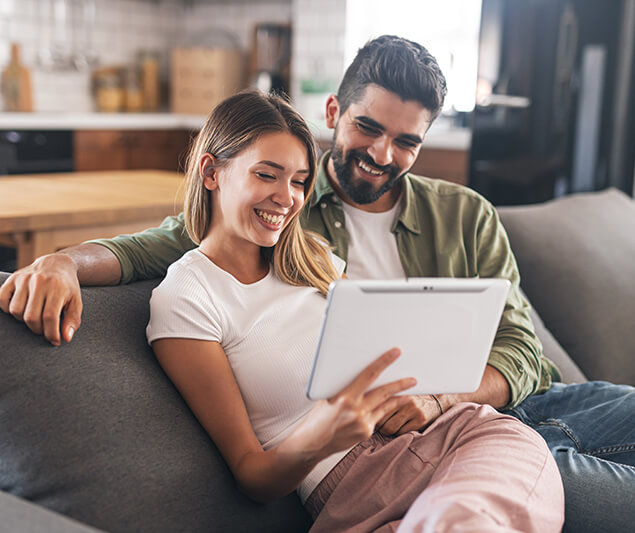 Imagen redonda de una pareja joven sonriente sentada en el sofá mirando tablet y la cocina está en segundo plano.