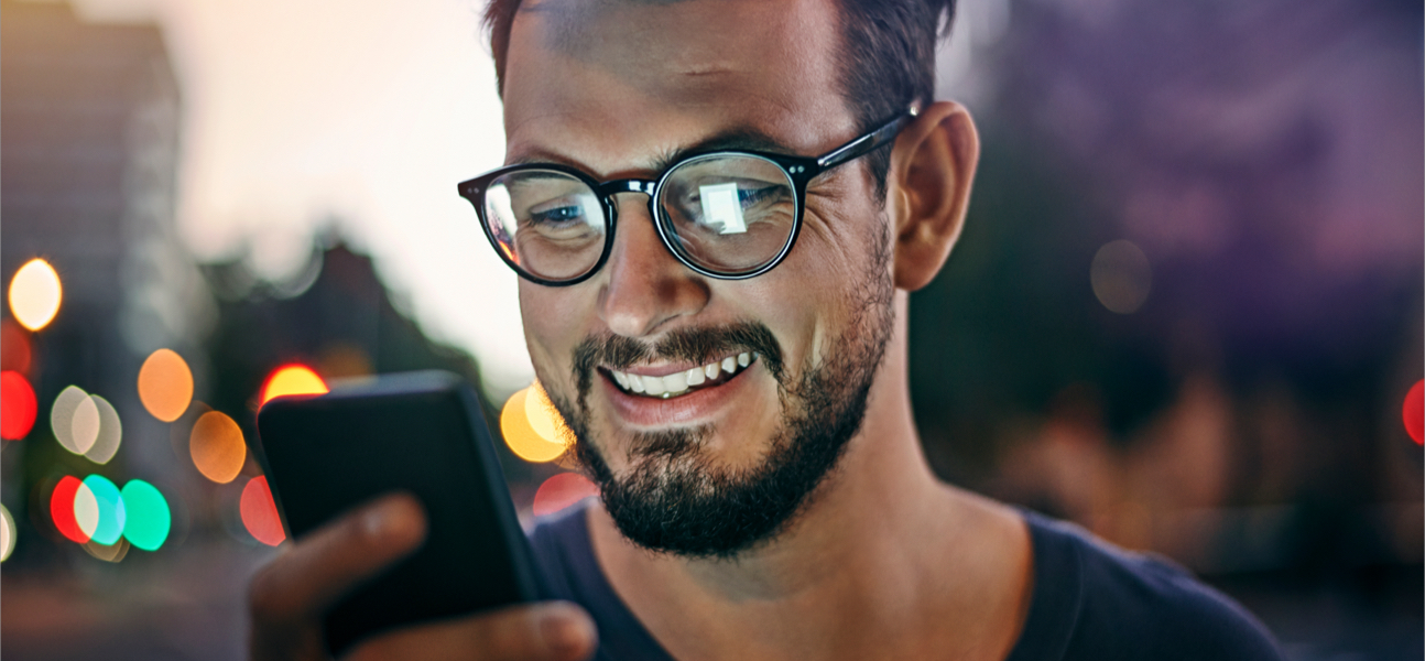 Man with beard and glasses sitting outside smiling looking at mobile phone