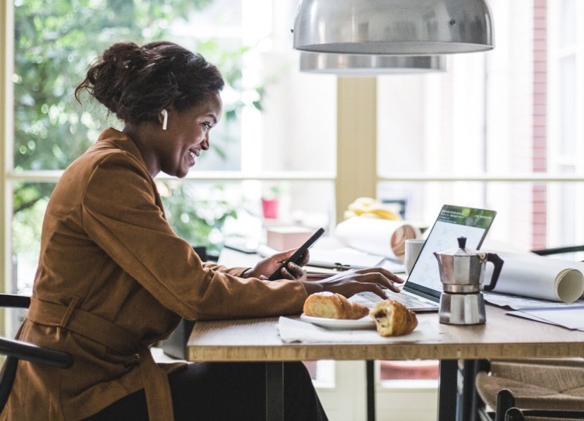 Mujer sonriendo sentada a la mesa con un café y plato mirando su laptop mientras sostiene el teléfono móvil