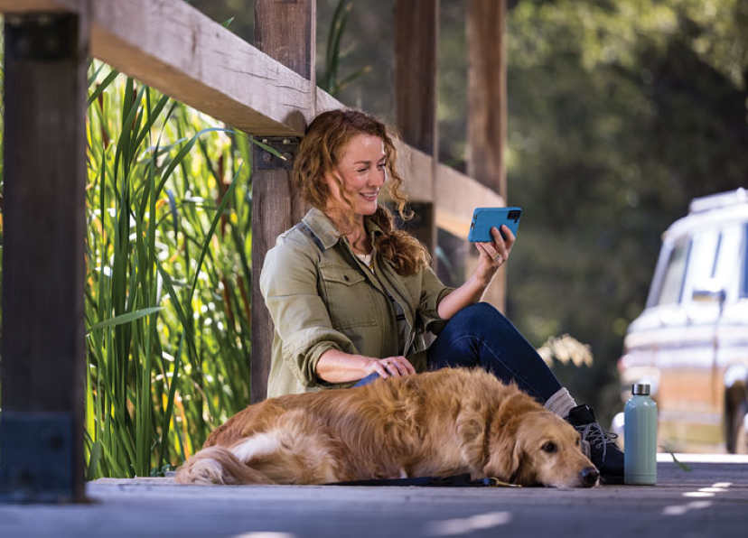 Mujer sonriendo con un perro, sentados al aire libre sobre un puente, luego de caminar sosteniendo un teléfono móvil